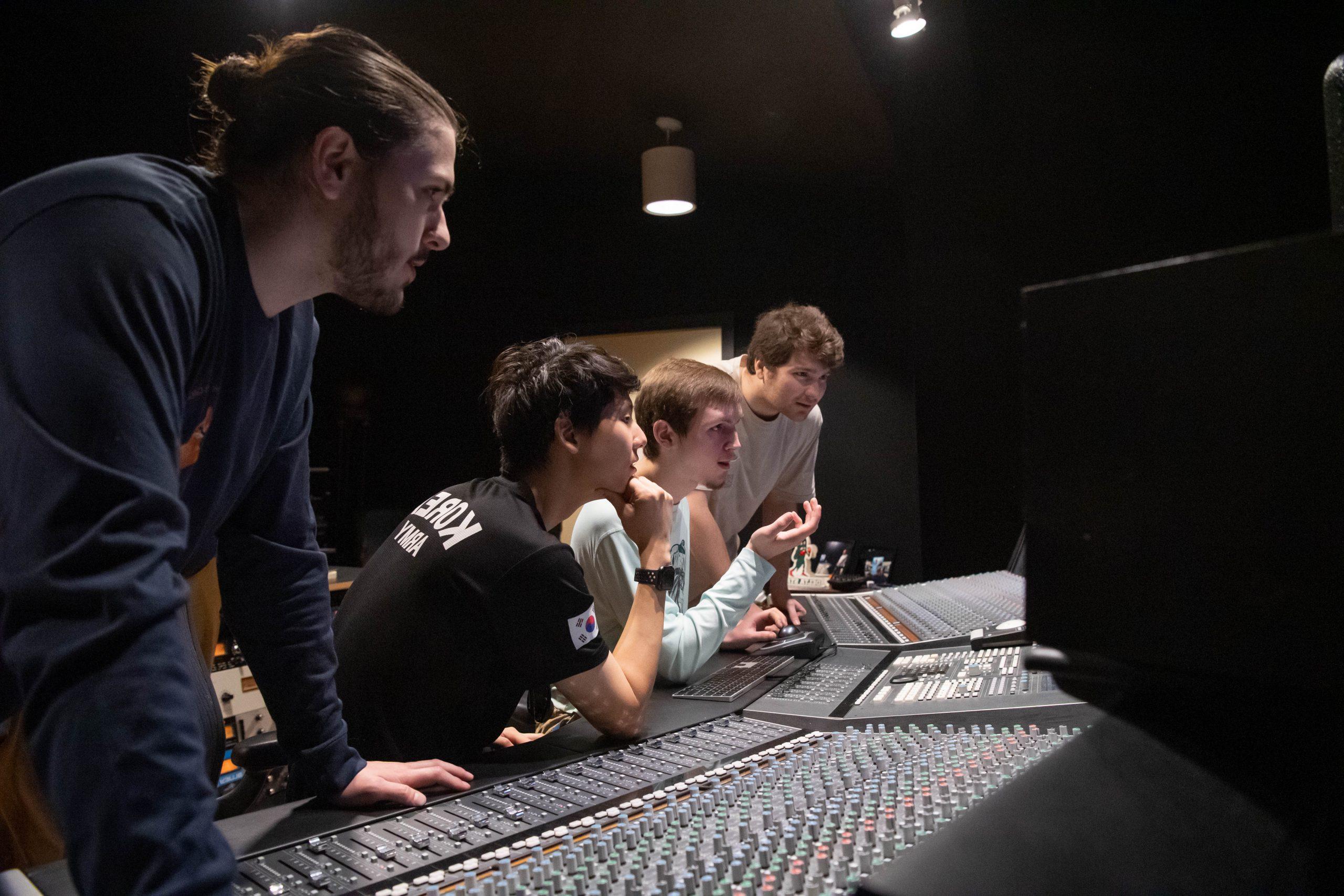 Four students standing and sitting in front of recording studio console, working on a project.