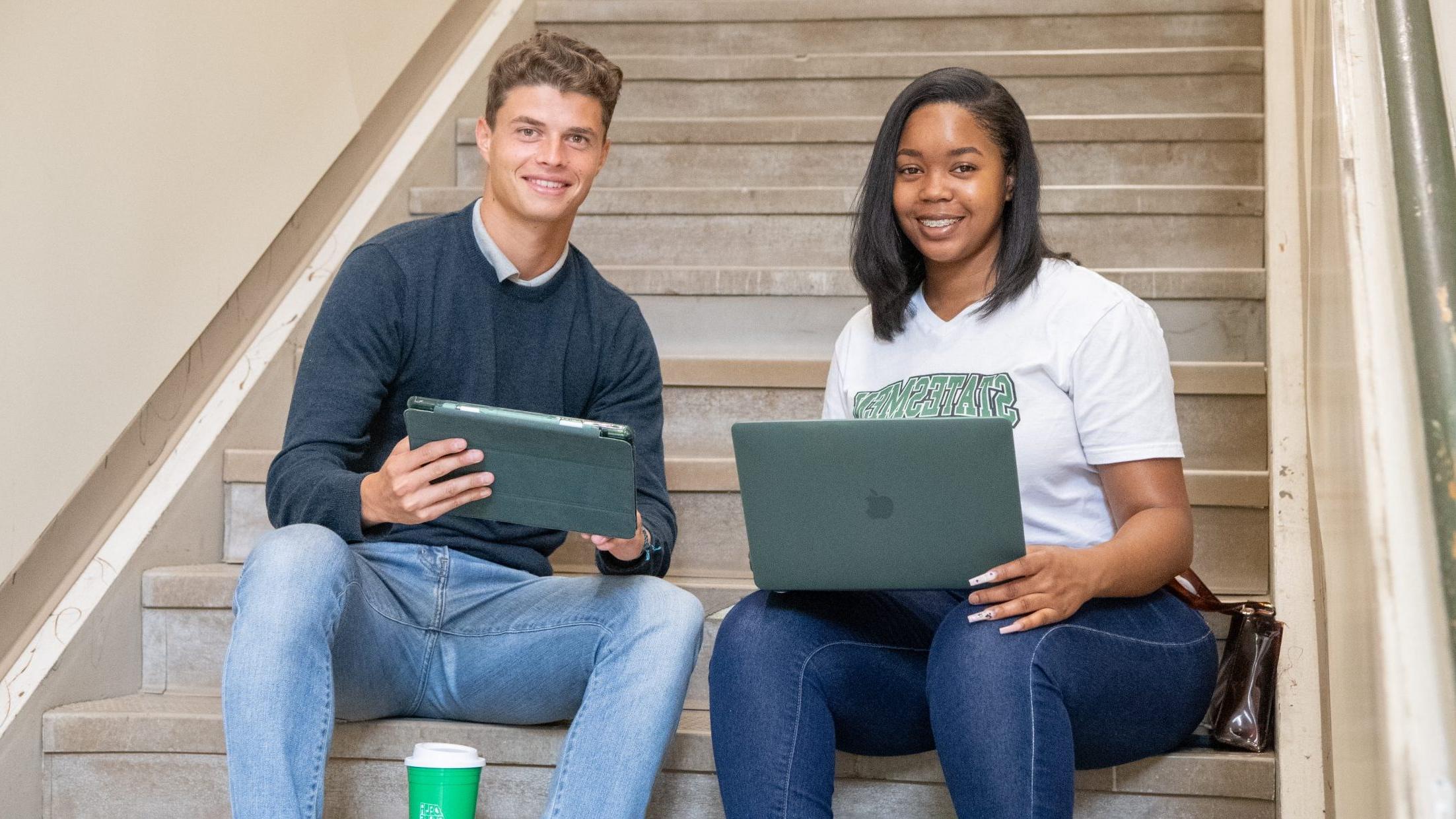 Two students, one with laptop and one with an ipad, sitting on stairs smiling.