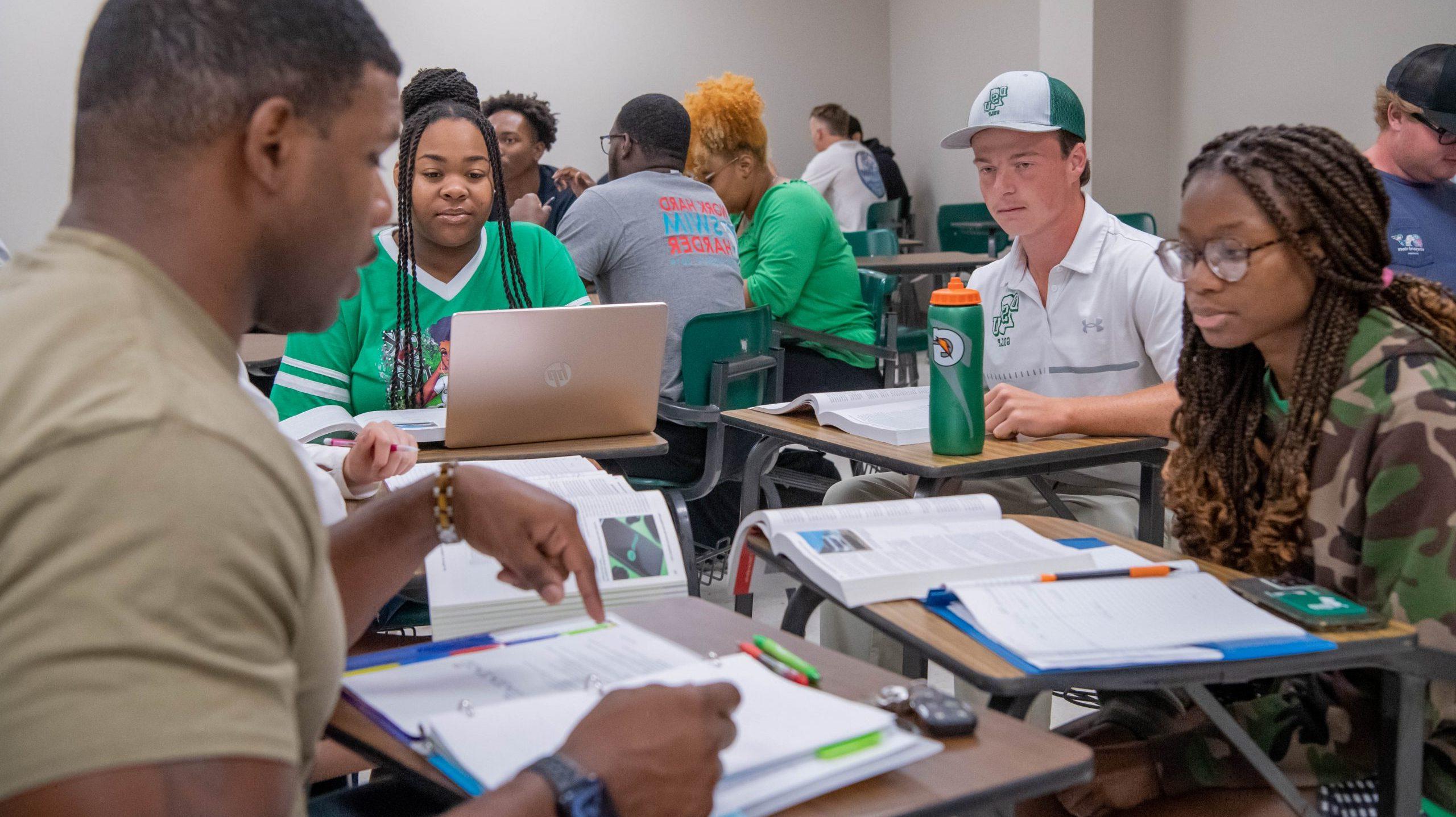 A group of four students working together on an assignment with other students in the background.